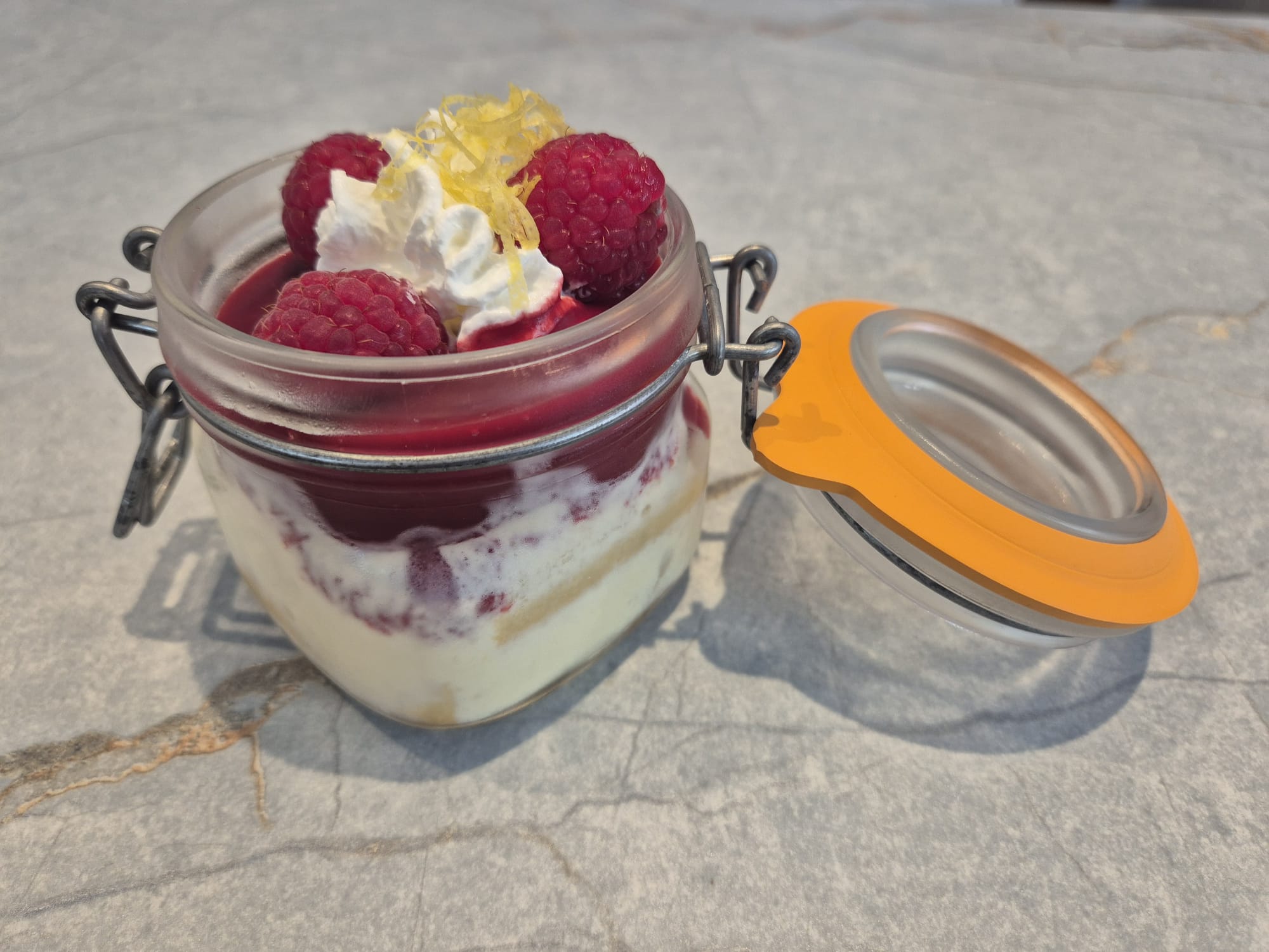 Glass of Limoncello Tiramisu topped with raspberry coulis and fresh raspberries, served with a lemon slice on the side.